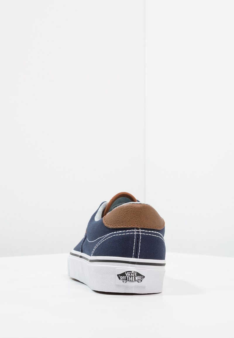 Zapatilla de lona navy con acento de piel marrón en el talón. Suela blanca con rayas negras y logo de Vans en el lado exterior. Detalle de costura visible.