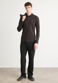Langærmet brun polo med krave og logo på brystet, parret med sorte bukser og sorte snikere, mens der holdes en mørk sok.