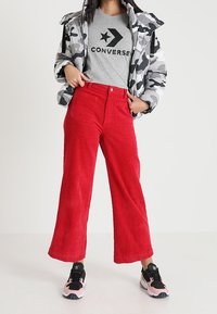 Czerwone szerokie spodnie z korduroy z przednimi kieszeniami, w połączeniu z szaro-camuflażową kurtką puchową oraz szarym t-shirtem z czarnym logo, a także czarnymi trampkami.