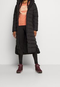 Persona che indossa un lungo piumino nero, leggings neri, scarponi da trekking color marrone scuro e una maglietta arancione con la scritta "soil power".