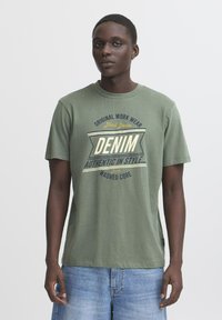 Zelené bavlněné tričko s krátkým rukávem a kulatým výstřihem, s potištěným grafickým prvkem, který zahrnuje slovo "DENIM" a dekorativní prvky.