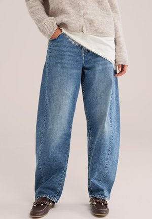 Personne portant un jean bleu large, un pull beige, un haut blanc bordé de dentelle et des mocassins marron foncé, debout avec une main dans la poche.