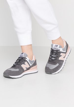 Para szarych i różowych sneakersów New Balance 574 noszonych z białymi spodniami na jasnym tle, stopy w pozycji pół kroku.