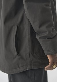 Schwarze Jacke mit einer seitlichen Tasche mit Reißverschluss, aus einem glatten, robusten Stoff gefertigt. Das Design umfasst einen geraden Saum und eine strukturierte Oberfläche.