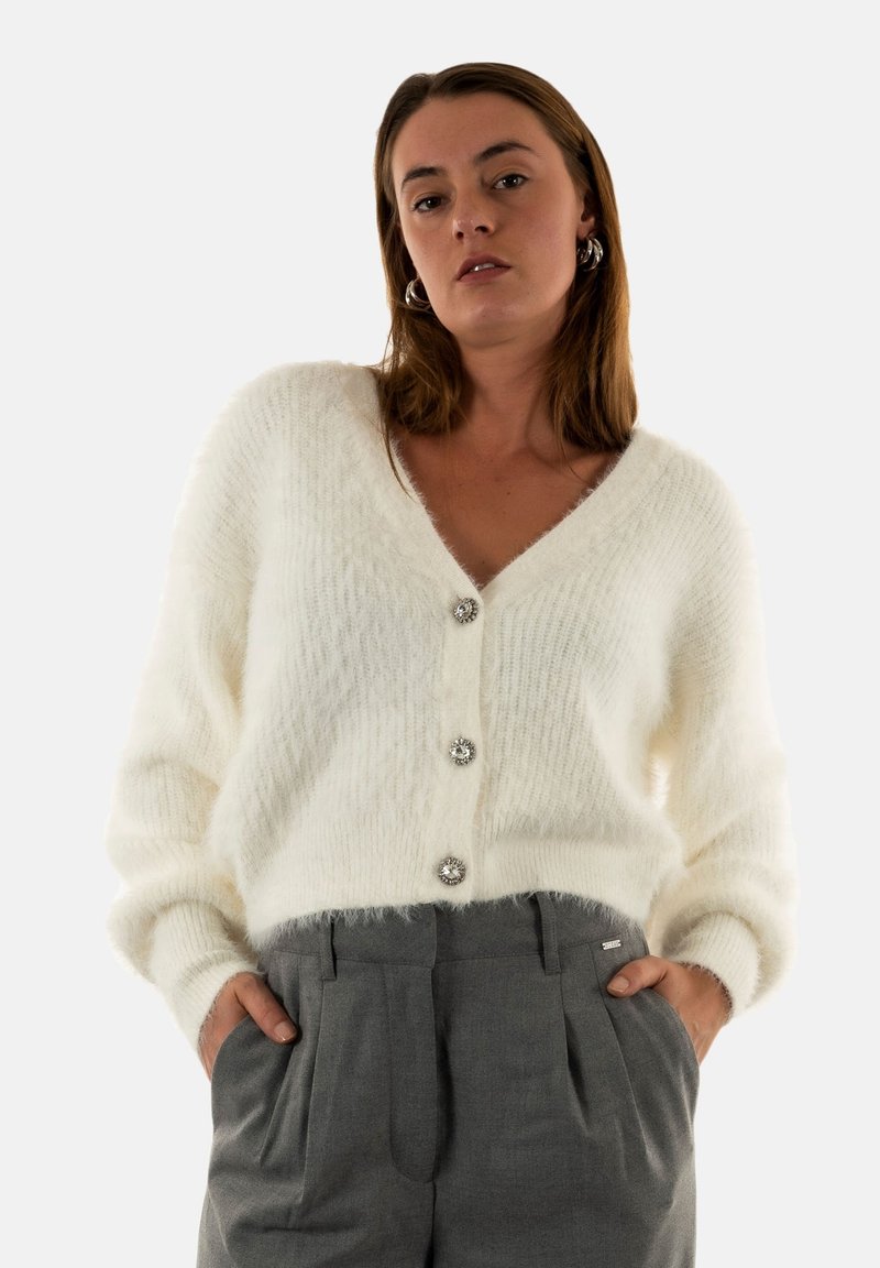 Cardigan blanc duveteux avec un col en V, orné de trois boutons décoratifs et de poignets côtelés. Porté par-dessus un pantalon gris taille haute.