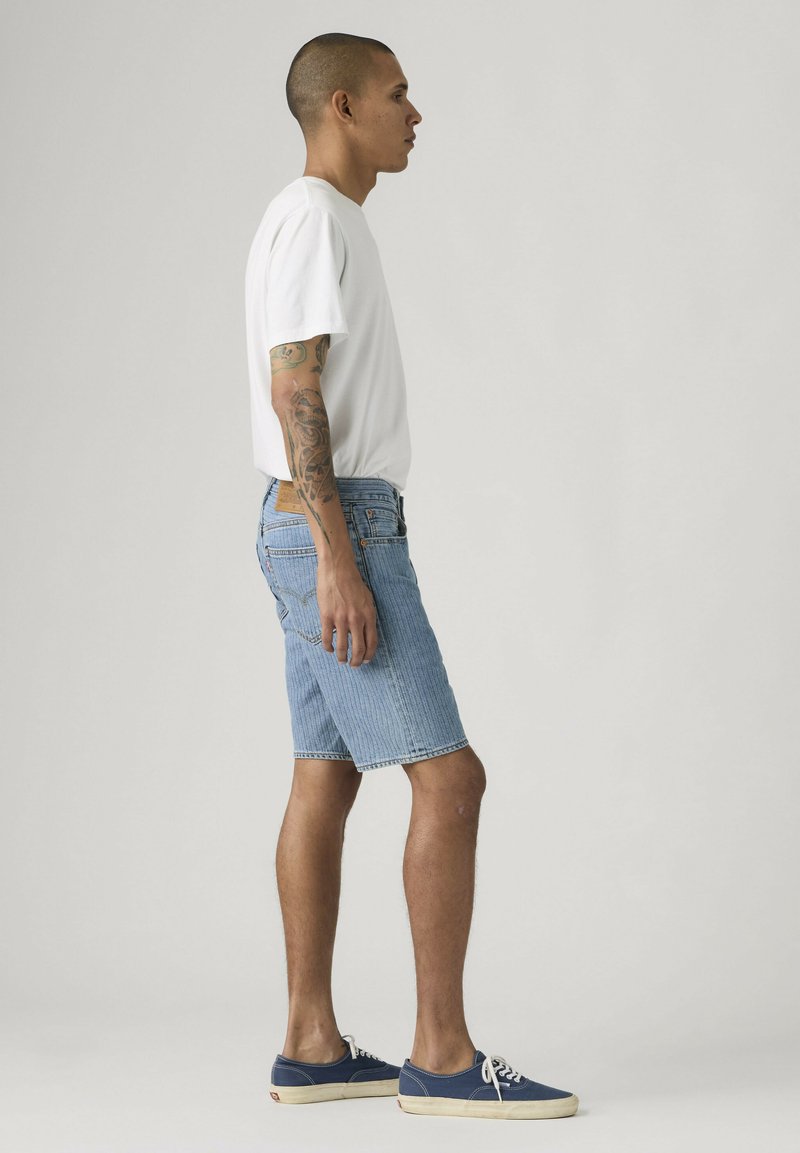 Hombre con brazo tatuado que lleva camiseta blanca, pantalones cortos de mezclilla y zapatillas azules, está de perfil contra un fondo simple.