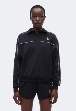 Personne portant une chemise noire à manches longues et un short noir avec un liseré blanc et de petits logos carrés colorés sur la poitrine et le short.