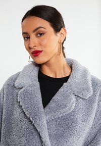 Grå faux pels frakke med en tekstureret, fluffy overflade, skrå krave og frontknapper. Modellen har en sort top på under.