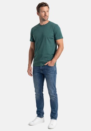 Mand stående med den ene hånd i lommen iført mørkegrøn kortærmet t-shirt, blå jeans og hvide sneakers mod en hvid baggrund.