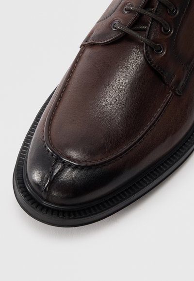 Zapato de cuero marrón con textura suave, punta redondeada y costuras en contraste. Suela de goma negra con detalle de borde acanalado.