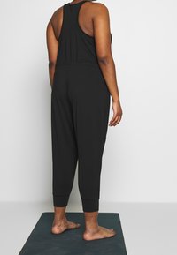 Salopette noire avec un design sans manches, taille cintrée et poignets fuselés. Texture lisse et tissu léger.