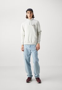 Ανοιχτό γκρί φούτερ με μπροστινή τσέπη και κεντημένο λογότυπο, σε συνδυασμό με ανοιχτό μπλε τζιν παντελόνι και μπορντό αθλητικά παπούτσια. Μαλακή υφή, άνετη γραμμή.