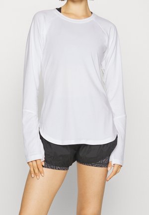 Person mit weißem langärmeligen Sportshirt und schwarzen Shorts, steht vor einem schlichten hellen Hintergrund, von den Schultern bis Mitte Oberschenkel sichtbar.