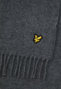 Šedý vlnený šál s třásněmi na koncoch a malým žltým vyšitým logom vtáka pri jednom okraji.