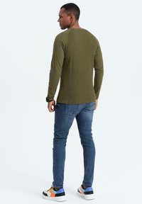 Camisa de manga larga color verde oliva con textura acanalada, combinada con jeans azules y zapatillas multicolor. Vista desde la parte trasera.