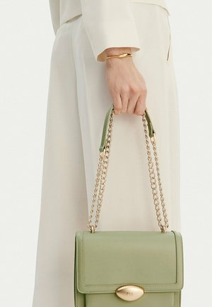 Ruka držící světle zelenou kabelku se zlatými řetízkovými popruhy, nošenou s krémovými kalhotami a zlatým náramkem na zápěstí.