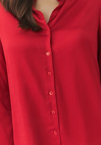 Red button-up shirt made of smooth fabric, featuring matching buttons and a simple collar design. No patterns or embellishments visible.
