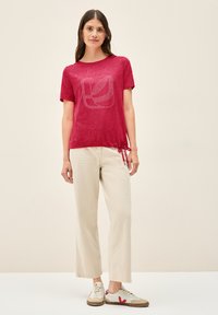 Rød t-shirt med korte ærmer og bladmønster, med en snørelukning i kanten, parret med beige brede bukser og hvide sneakers.