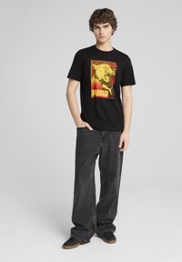 Camiseta de algodón negra con un estampado gráfico en amarillo y rojo que presenta un león y el texto "PUMA", combinada con unos jeans oscuros de corte holgado y zapatos negros.
