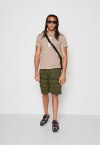 Beige kortærmet t-shirt med logo, olivengrøn cargo shorts og mørke sneakers med farverige accenter. Slim fit, tekstureret stof, afslappet design.
