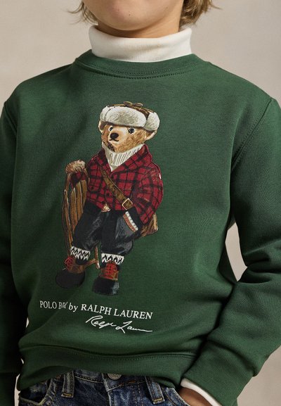 Grønn genser med en bjørneillustrasjon som har på seg en rød ruterjakke, svarte bukser og snøstøvler, med teksten "POLO by RALPH LAUREN".