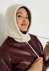 Mujer con capucha de punto acanalado color crema sujetando los cordones, vestida con una chaqueta de cuero color burdeos oscuro, contra un fondo claro y liso.