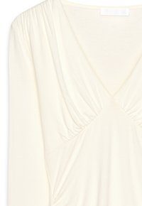 Top léger crème à manches longues avec un col en V et des détails froncés aux épaules, en tissu doux. Pas de motifs ou d'embellissements visibles.