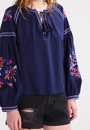 Vrouw draagt een marineblauwe blouse met geborduurde bloemmotieven op de mouwen en zwarte gescheurde denimshorts, staand tegen een effen achtergrond.