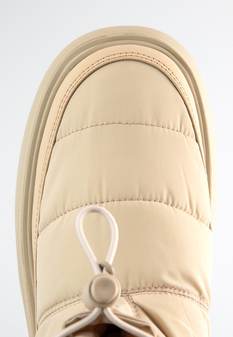 Beige isolierter Winterstiefel mit gesteppter Oberseite, runder Zehenpartie und Schnürsystem mit Zugverschluss. Dicke Gummisohle mit strukturiertem Profil.