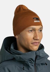 Brown ribbed beanie with a pointed top and a logo patch featuring mountains and the brand name "BUFF," worn with a gray jacket.