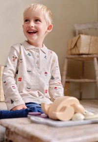 Světlé béžové tričko s dlouhým rukávem a různými barevnými vánočními výšivkami, v kombinaci s modrými džíny. Dítě sedí na dřevěné podlaze.
