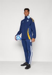 Mornarska sportska jakna s žutim naglascima, bijele pruge na tamnoplavim hlačama, crni kopački i šarena nogometna lopta.