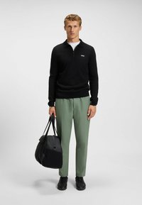 Sorte lynlås-pullover, grønne bukser og en sort duffelbag. Sorte sko fuldender looket. Enkel design med minimal branding.