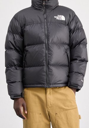 Černá péřová bunda s předním zipem a bílým logem "The North Face" na hrudi, nošená přes béžové kalhoty. Ruce částečně viditelné.