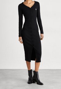 Pitkät hihat, musta ribbineulemekko, jossa on V-pääntie ja etupainike. Sisältää sivureiän ja pienen logotunnuksen rinnassa.