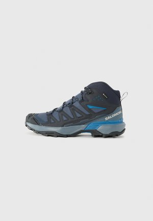 Scarpa da trekking outdoor di altezza media in blu scuro e grigio, dotata di suola robusta, tessuto superiore strutturato e dettagli riflettenti. Design leggero.