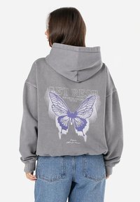 Pilkas didelis hoodie su dideliu purpuriniu drugelio piešiniu ir tekstu ant nugaros. Pagamintas iš minkštos medžiagos, turi kengūrinę kišenę.