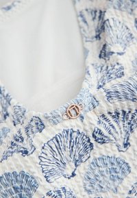 Gros plan d’un tissu blanc avec motif de coquillages bleus et petite boucle ronde dorée près de l’encolure froncée.