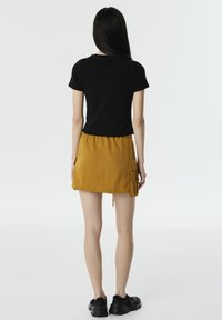 Zwarte korte mouwen t-shirt gecombineerd met een mosterdgele rok met een zijzak en een trekkoord in de taille. Draagt zwarte sneakers.