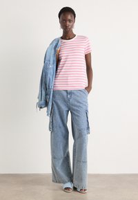 Stripete rosa og hvite bomull T-skjorte, kombinert med lyseblå denim cargo-bukser og en denimjakke som henger over skulderen. Blå sandaler.