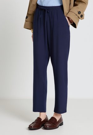 Persona che indossa pantaloni blu con coulisse, mocassini marroni, una camicia azzurro chiaro e una giacca color sabbia con le mani in tasca.