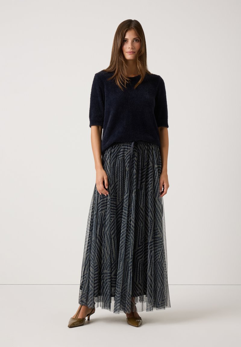 Navyblauer, fuzzie Kurzarm-Pullover kombiniert mit einem langen, gemusterten Tüllrock in Marineblau und Weiß. Beigefarbene Absatzschuhe vervollständigen das Outfit.