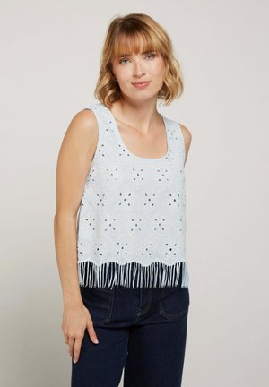 Femme aux cheveux blonds courts portant un haut sans manches blanc en œillet avec des motifs floraux et des franges, associé à un jean bleu foncé.