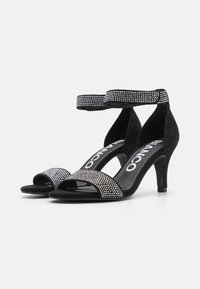 Sandali con tacco alto in suede nero, dettagli in strass, cinturino alla caviglia, design open toe e tacco sottile. Nome del marchio visibile sulla soletta.