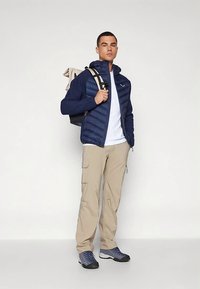 Mørkeblå pufferjakke med tætsiddende design over en hvid t-shirt, beige cargobukser og grå vandresko. Rygsæk med beige og sorte stropper.