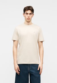 Νέος άνδρας με κοντά καστανά μαλλιά φοράει ένα απλό μπεζ κοντομάνικο μπλουζάκι και σκούρα μπλε τζιν, στέκεται μπροστά σε ανοιχτό γκρι φόντο.