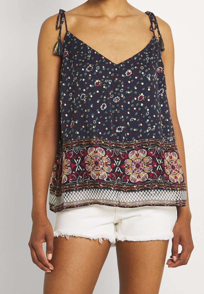 Personne portant un top sombre à fleurs sans manches avec des bretelles à nouer et un ourlet à motifs, assorti à un short en jean blanc effiloché, debout sur fond uni.