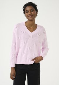 Pull en tricot rose clair avec un col en V, présentant un motif géométrique texturé et des manches longues. Associé à un jean noir.