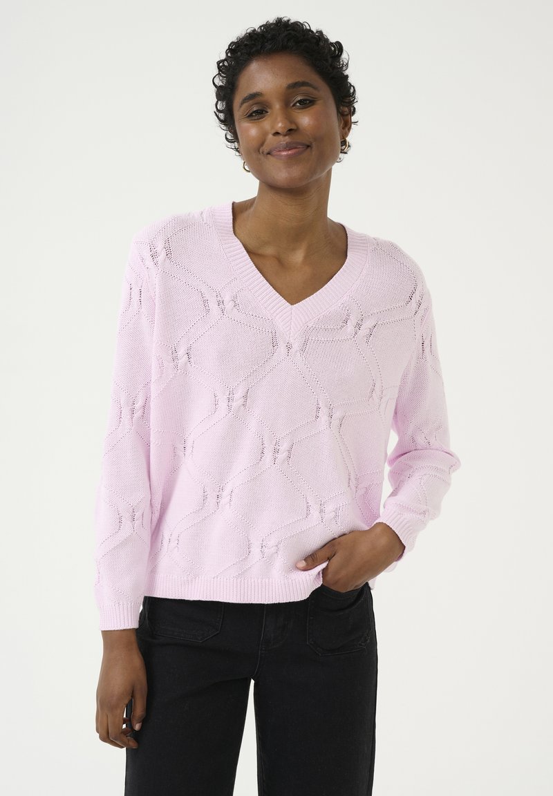 Pull en tricot rose clair avec un col en V, présentant un motif géométrique texturé et des manches longues. Associé à un jean noir.