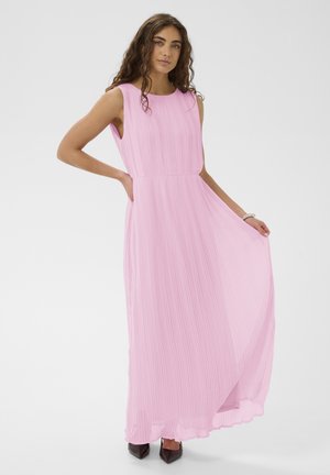 Femme portant une robe rose plissée sans manches, longue jusqu'au sol, tenant l'ourlet de la jupe d'une main, debout devant un fond blanc uni.